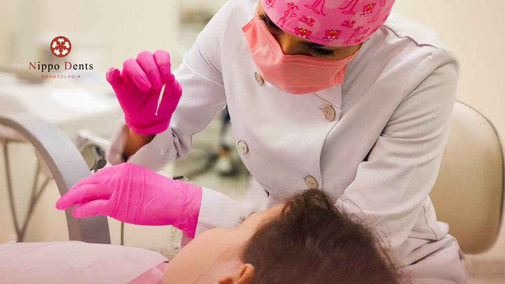 Dentista realizando atendimento infantil com luvas e máscara rosas, aproximando instrumento odontológico da criança deitada na cadeira